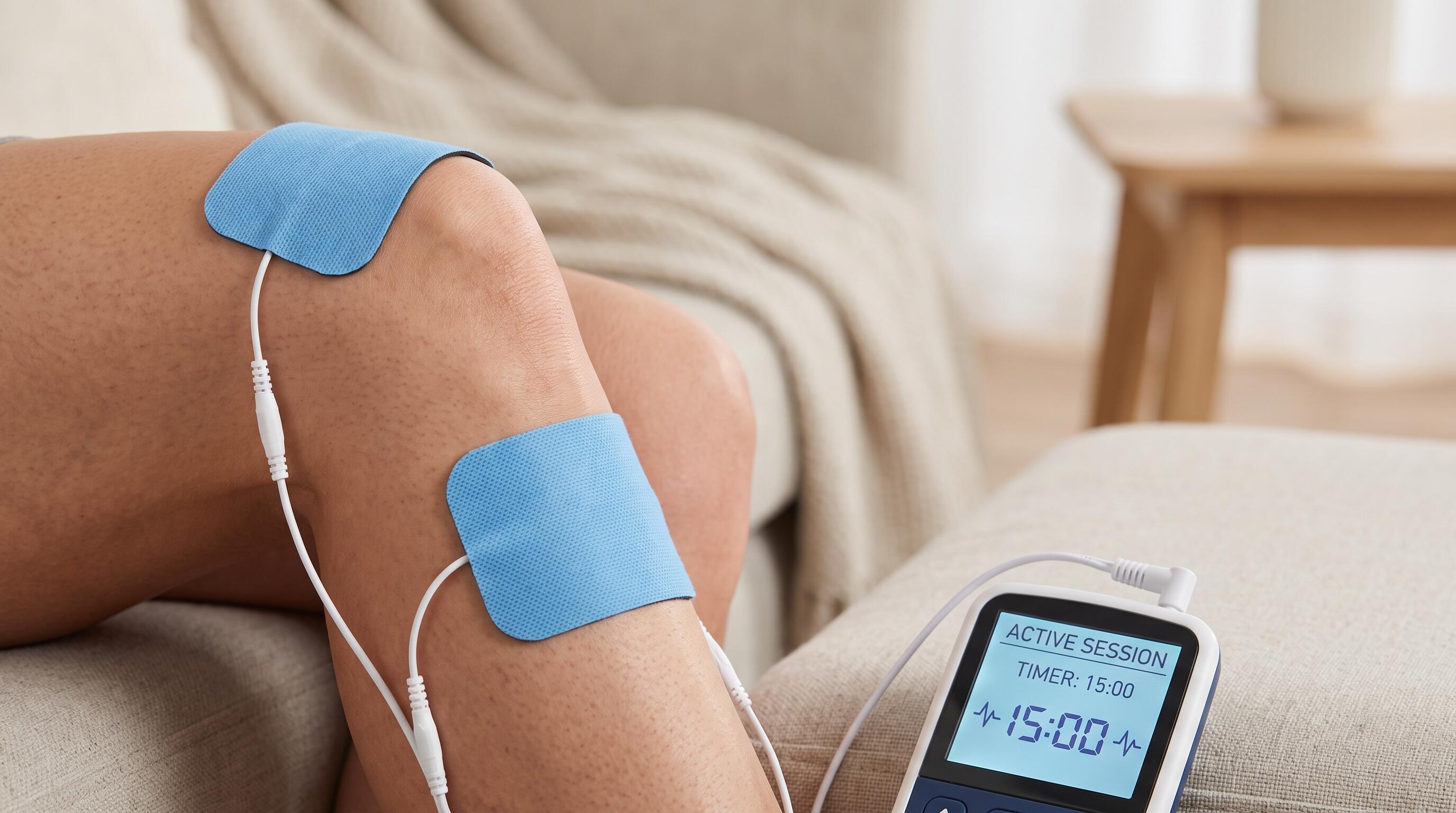 Close-up of TENS unit electrode placement on knee with device display showing active therapy session